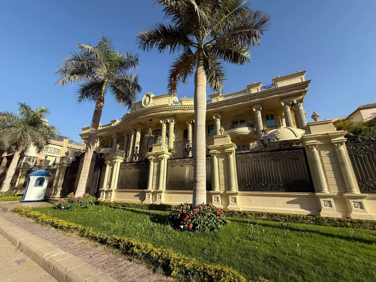 Exterior photograph of Gamal Hakam Villa in New Cairo showing its columned curved frontage, decorative gates and palm-lined garden edge